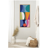 Vertical abstract canvas with colorful geometric shapes in blue, green, orange, purple, and pink framed in light wood, hanging on a pale gray wall above a beige carpet.