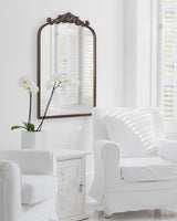Traditional arched wall mirror with carved dark wood frame above a white side table holding a white orchid, beside a white upholstered armchair in bright room.
