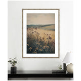 Framed art print depicting a wildflower meadow with tall grasses against soft hills and a pale sky in muted earthy tones, hanging above a black table with plants and a lamp.