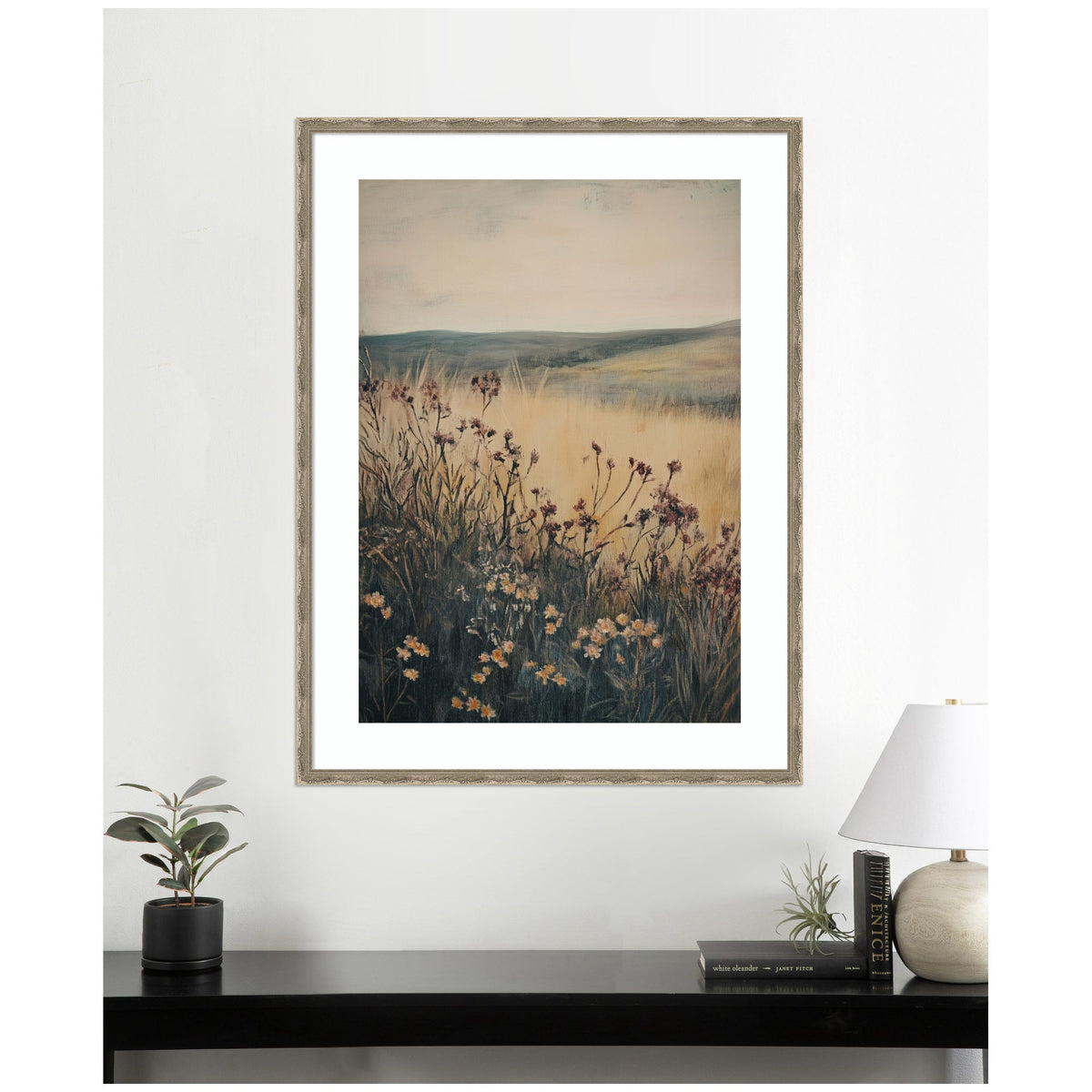 Framed art print depicting a wildflower meadow with tall grasses against soft hills and a pale sky in muted earthy tones, hanging above a black table with plants and a lamp.