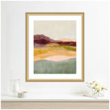 Abstract landscape art with layered shapes in muted green, pink, yellow, and brown, framed in light wood and displayed on a white wall above a shelf with flowers and candles.