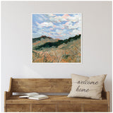 Framed canvas print of a landscape with green hills under a swirling blue and white cloudy sky above a tan grass field, hanging above a rustic wooden bench with a beige 'welcome home' pillow.