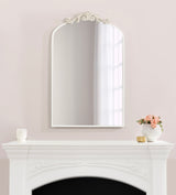 Arched wall mirror with a white decorative frame featuring floral and scroll details at the top, hanging above an ornate white mantel with floral accents.