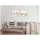 Two framed abstract landscape prints with soft beige, green, and gray tones hung above a beige sofa adorned with patterned black-and-white pillows and a chunky knit throw.