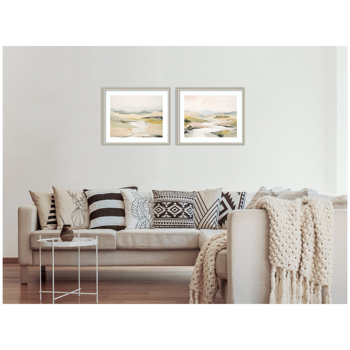 Two framed abstract landscape prints with soft beige, green, and gray tones hung above a beige sofa adorned with patterned black-and-white pillows and a chunky knit throw.