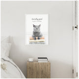 Framed canvas showing a gray cat resting on a stack of fluffy folded clothes with text 'laundry goal fluffy clothes' on a white wall above a wooden bedside table in a minimalist bedroom.