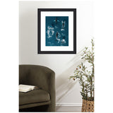 Framed aerial artwork of a school of humpback whales swimming in deep blue ocean water, displayed on a white wall above an olive-green armchair and beside a tall potted plant.