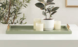 Rectangular muted green decorative tray with gold handles holding a white ceramic pot with a dark green leafy plant and four cream-colored candles on a white surface.