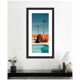 Framed vertical photo showing a bison lying by a turquoise pool with two tall palm trees and an orange wall in the background under a clear blue sky.