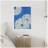 Framed canvas art featuring three white sand dollar sea shells with blue dot patterns on a textured cobalt blue background, hanging above a wooden bedside table.