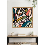 Square framed canvas wall art with abstract, colorful, overlapping leaf shapes in bold black outlines against a soft pink background, displayed above a wooden console table with neutral decor.