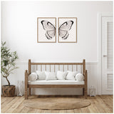 Two framed canvas prints displaying watercolor butterfly wings in neutral tones with linen texture, hung side by side above a wooden bench with white cushions in a minimalist room.