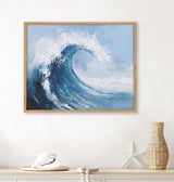 Abstract coastal painting showing a dynamic ocean wave curling with white foam against a light blue sky, framed in natural wood and hung above a beige shelf with seashell and starfish decor.