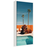 Framed vertical canvas showing a bison resting near a turquoise swimming pool with an orange wall and two tall palm trees against a clear blue sky.