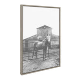 Black and white photograph of a horse standing behind a metal fence in front of a weathered wooden barn with a pitched roof, framed in a light wood frame.