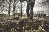 Person wearing jeans and boots digging soil with a shovel in a wooded area, with a Big Tine Nitro granular mineral supplement bag nearby and an off-road vehicle in the background.