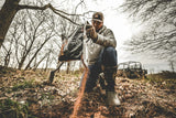 Person wearing boots and a cap pours granular mineral supplement from a black bag onto soil in a leaf-covered outdoor area with bare trees.