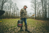 Man in camouflage jacket and boots holding a shovel and a black bag labeled Big Tine Nitro, standing on a grassy, leaf-strewn path surrounded by bare trees.
