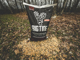 Black Big Tine Fortified Deer Blend Cherry bag with white deer logo standing upright outdoors on leaf-covered ground with feed spilled in front beneath bare winter trees.