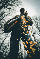 Person wearing dark clothing uses a black leaf blower labeled 'WATER DEER BLEND NITRO' to blow orange and beige debris outdoors among bare trees.