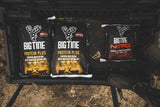 Three bags of Big Tine deer feed, two labeled Protein Plus with black and yellow designs, and one labeled Nitro with black and red details, placed on a vehicle tailgate.