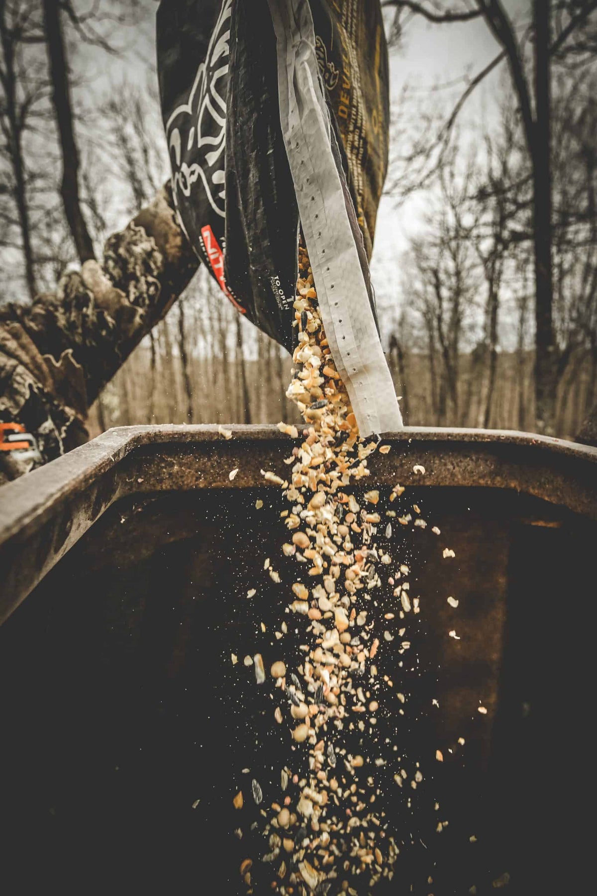 Wildlife feed corn and grains falling from a partially opened black and beige bag into a metal container outdoors with leafless trees in the background.
