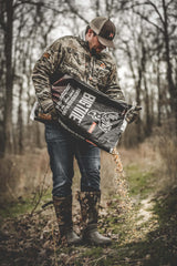 Man in camouflage jacket and boots pouring Big Tine fortified deer blend cherry bait onto a forest floor covered with leaves and grass.