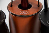 Close-up of round brown leather wine tote top with stitched edges and engraved logo, holding a wine bottle neck, flanked by black leather totes.