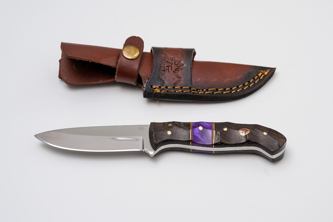 Hand-Forged Satin Finish Hunter knife with purple pearl resin and dark textured wood hybrid handle lying next to a brown leather sheath