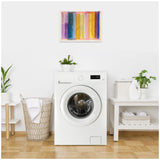 Front-loading white washing machine in a bright laundry room with a woven laundry basket, two potted green plants, and colorful abstract artwork mounted above the machine.
