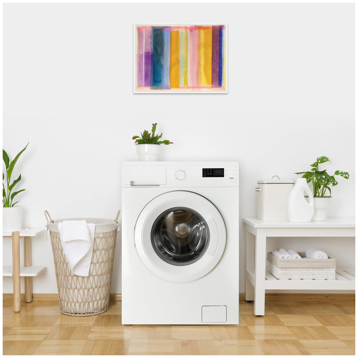 Front-loading white washing machine in a bright laundry room with a woven laundry basket, two potted green plants, and colorful abstract artwork mounted above the machine.
