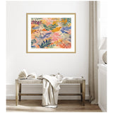 Abstract botanical artwork with overlapping leaves in warm pink, orange, blue, and green hues, framed in light wood and hung above a rustic wooden bench with a draped cream throw.