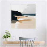 Abstract coastal landscape painting with sandy beige shore, dark cliffs on the left, and muted gray sky above, framed and hanging above a light wooden desk.