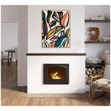 Framed abstract botanical canvas with colorful stylized leaves in teal, pink, yellow, and black, hung above a white brick fireplace with a burning wood stove insert.