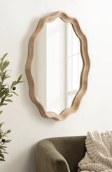 Round scalloped mirror with a natural wood frame, reflecting a window, hung on a white wall above an olive green chair with a chunky knit blanket.