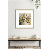 Square framed print of a branch with green olives and leaves in soft muted tones, displayed on a white wall above a wooden table with books and decor.