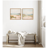 Two framed square canvas paintings of soft pastel landscapes with sandy dunes and distant blue water, hanging side by side on a white wall above a wooden bench with a beige throw and open book.