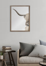 Vertical framed print of a close-up Highland cow portrait showing its long curved horn and shaggy hair against a plain background, hung above a neutral-toned sofa.