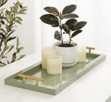 Rectangular sage green tray with gold handles holding a small potted plant with dark green leaves and three ivory candle holders, set on a white surface.