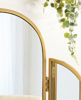 Close-up of a gold metal tri-fold arch mirror with one folded panel and green hanging plant leaves in the background on a white wall.