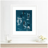 Framed aerial view of a school of humpback whales swimming in deep blue ocean, with water swirling around their bodies, displayed on a white wall above a light wood shelf.
