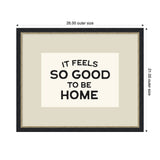 Framed wall art with black bold typography reading 'It feels so good to be home' centered on a light beige background with a dark, ornate border and gold beaded inner trim.