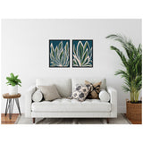 Two framed canvases with abstract white, green, and beige leaf shapes on deep blue backgrounds hung side by side above a white sofa with patterned pillows.