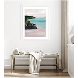 Framed seascape artwork showing a turquoise ocean with dark rocks and green foliage along a coastline under a cloudy sky, hung above a wooden bench with a blanket and open book.