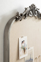 Close-up of an ornate gray arched frame pinboard with beige fabric backing, decorated with carved floral motifs and holding two pinned photographs.