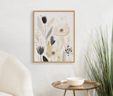 Framed floral print with muted beige, gray, black, and gold flowers and leaves, hung on a white wall above a brass side table with a cream bowl and green tall grass plant