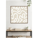 Square framed canvas print with beige background and white abstract organic curving lines, hanging above a wooden shelf with books, a cup, and a small white vase with greenery.