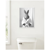 Black and white framed portrait of a rabbit wearing a white bathrobe, hanging above a white toilet against a plain white wall.