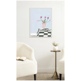 Framed canvas print of pink tulips in a pale pink vase on a black-and-white checkered table against a light blue background, hanging above two white armchairs with a brass side table between them.