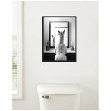 Black-and-white framed art print depicting a llama facing a mirror that reflects its face, hung above a white toilet with two small potted plants on the sink counter.